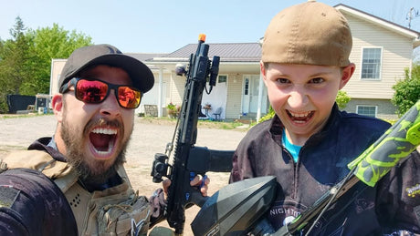 Nightmare PB posing with his son, holding an EMF200 magfed paintball marker, both smiling in paintball gear.